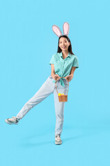 Young Asian woman in bunny ears with basket of Easter eggs on blue background