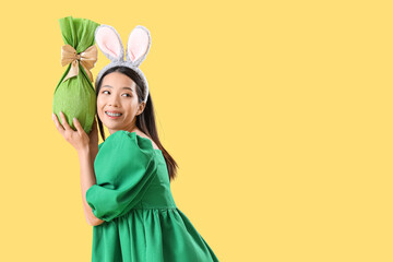 Young Asian woman in bunny ears with Easter gift egg on yellow background - Powered by Adobe