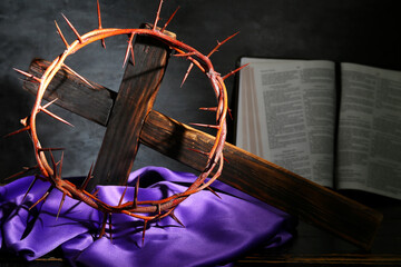 Wooden cross with crown of thorns, Bible and purple cloth on grey grunge background