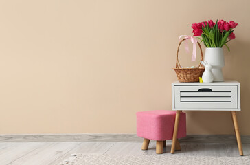 Vase with tulips and basket of Easter eggs on table near beige wall in room