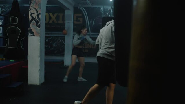 Athletic fighter in boxing bandages prepares for fighting championship and trains in dark boxing gym. Male boxer hits punching bag and practices fighting technique. Physical activity and workout.