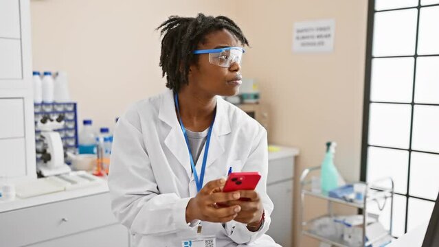 Confident young black woman with dreadlocks expressing serious attitude while working in lab, deep in thought over smartphone