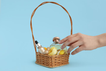 Female hands with wicker basket full of decorative cosmetics, Easter eggs and flowers on blue background