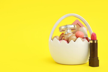 Basket in shape of eggshell with decorative cosmetics and Easter eggs on yellow background