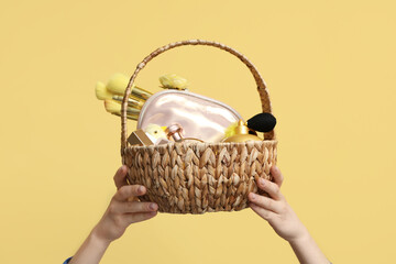 Female hands with wicker Easter basket full of decorative cosmetics and baby chicken on yellow...