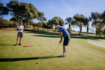 Obraz premium Sotogrante, Spain - January 25, 2024 - Two men on a golf green, one putting towards the hole, the other observing, with trees in the background.