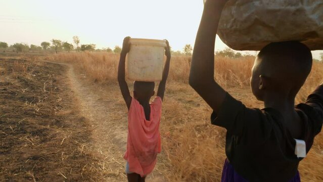 couple of black african child children kids carrying purified water in plastic bucket over their head at sunset walking in remote rural africa