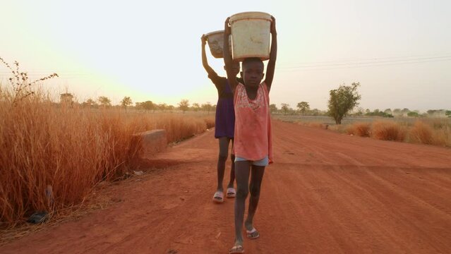 couple of african black child children kid walking in narrowed dusty road in remote village of africa with bucket over their head