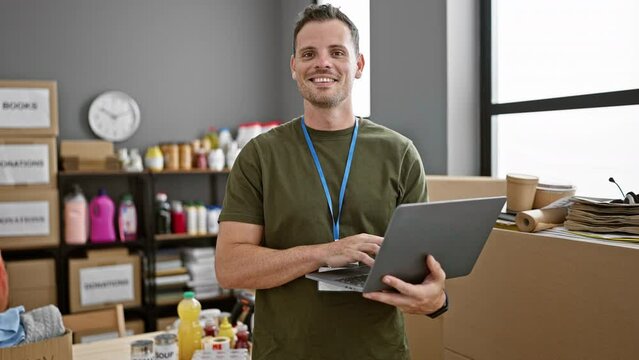 Smiling young hispanic man with a laptop stands in a warehouse workspace between cardboard boxes and supplies