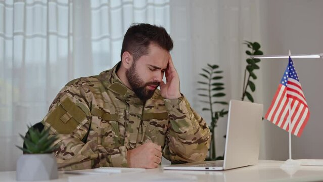 American veteran working by laptop while sitting by table with national flag and suffering from high blood pressure. Caucasian male wearing military uniform and getting distracted by sharp pain.
