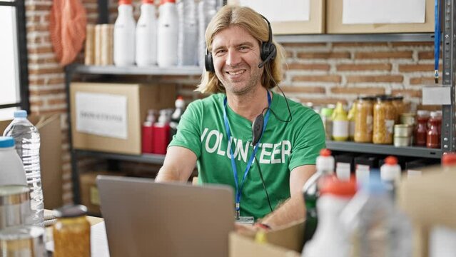 Long-haired man in green 'volunteer' shirt using a laptop in an indoor warehouse location, organizing donations.