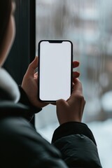 Mockup image of a woman's hands holding a smartphone with a white screen on the background of the window