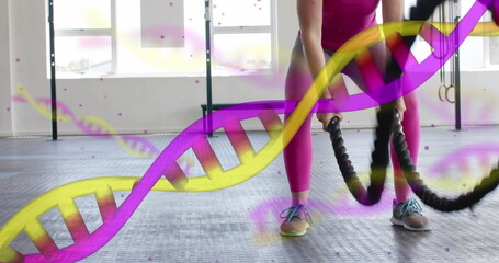 Image of dna strands over happy caucasian woman cross training with battle ropes at gym