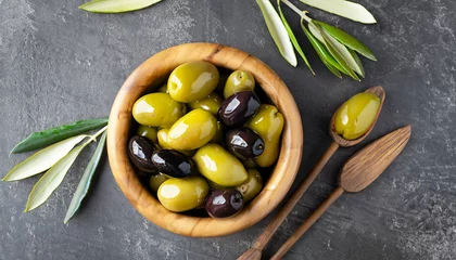 Fototapeten Fresh olives in wooden bowl on dark stone table. Black and green olive with pickers or sticks from top view. © Uuganbayar