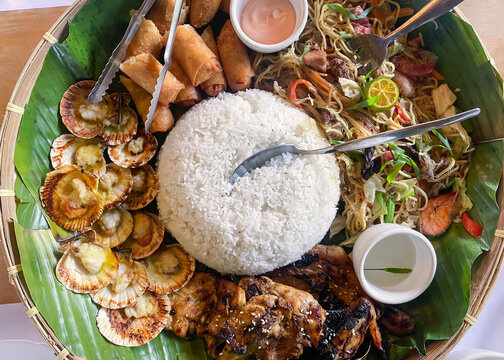 Filipino food on a plate with a banana leaf base. Rice, chicken, lumpia, pansit