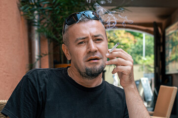 Handsome man smoking a cigarette in an outdoor cafe. Portrait of a young man in a black T-shirt with a cigarette in his hand