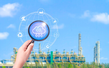 Hand holding magnifying glass with reduce CO2 emission icons on blurred oil industrial factory...
