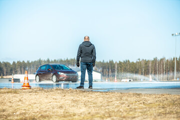 man observing a wet road test with a maroon car skidding on a water-covered track, testing vehicle...