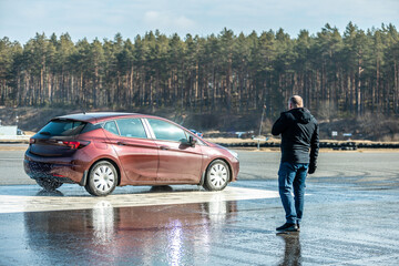 man observing a wet road test with a maroon car skidding on a water-covered track, testing vehicle...