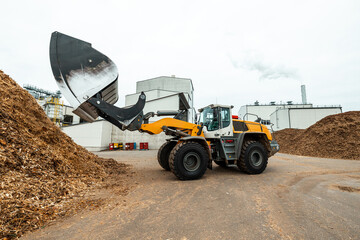 industrial wheel loader with raised bucket at a biomass power plant, handling wood chips against a...