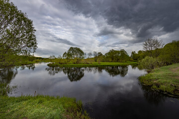 The river reflects the dramatic sky, a spring motif with the river and green bushes and grass.