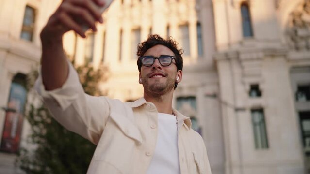 Cheerful young man blogger takes selfie with smartphone camera, sporting casual attire and glasses. Male tourist in wireless earbuds  photographing himself near historic building. Travel exploration
