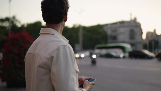 Young man waiting on street in early evening. Male looks intently at smartphone, monitoring arrival of taxi cab he ordered through app. City lights and passing traffic add dynamic urban energy 
