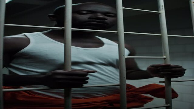 Vertical shot of African American inmate walking in prison cell, holding bars and looking at camera