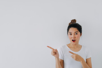 Asian Thai woman wow surprised face and pointing fingers to empty space, recommending products at blank background, standing isolated over white wall.