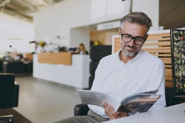 Male happy calm rich smiling successful businessman man wears classic shirt read catalog sitting at office table in dealership store indoors want to buy new automobile car. Business lifestyle concept. - obrazy, fototapety, plakaty