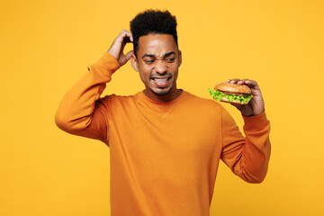 Young confused sad upset man wear orange sweatshirt casual clothes hold eat burger scratch head isolated on plain yellow background studio. Proper nutrition healthy fast food unhealthy choice concept.