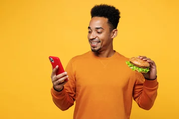 Foto op Plexiglas Young man wear orange sweatshirt casual clothes hold use mobile cell phone eat burger check calories isolated on plain yellow background. Proper nutrition healthy fast food unhealthy choice concept. © ViDi Studio