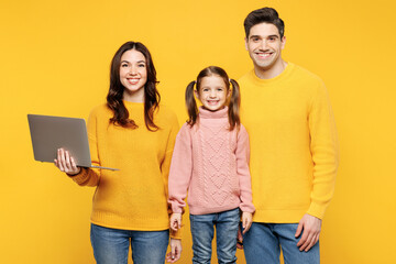 Fototapeta premium Young smiling happy IT parents mom dad with child kid girl 7-8 years old wear pink sweater casual clothes hold use work on laptop pc computer isolated on plain yellow background. Family day concept.