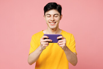 Young man he wears yellow t-shirt casual clothes use play racing app on mobile cell phone hold gadget smartphone for pc video games isolated on plain pastel light pink background. Lifestyle concept.