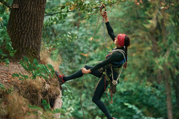 View from the side, hanging on the rope. Woman is doing climbing in the forest by the use of safety equipment
