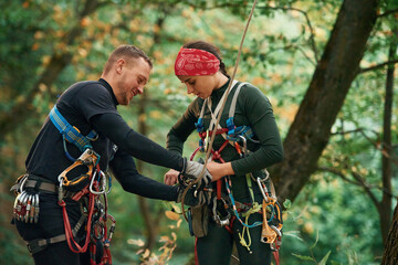 Man is helping the woman. Doing climbing in forest with use of safety equipment
