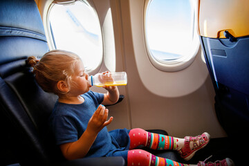 Adorable little girl traveling by an airplane. Small toddler child drinking orange juice sitting near aircraft window. Traveling abroad with kids. Family on summer vacations.