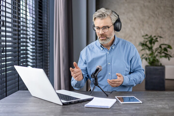 Focused aged male in headphones sitting by working table with laptop and professional microphone...