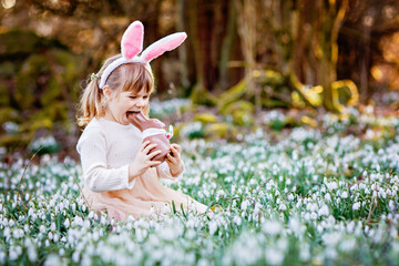 Happy little girl with Easter bunny ears eating chocolate figure in spring forest on sunny day, outdoors. Cute child with lots of snowdrop flowers. Springtime, christian holiday concept.
