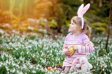 Little girl with Easter bunny ears making egg hunt in spring forest on sunny day, outdoors. Cute...