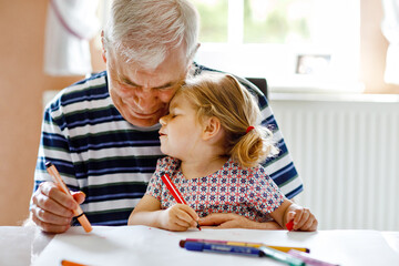 Cute little baby toddler girl and handsome senior grandfather painting with colorful pencils at...