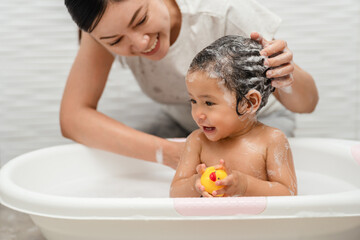 mother bathing and washing her infant baby hair with shampoo while playing a rubber duck toy in bathtub