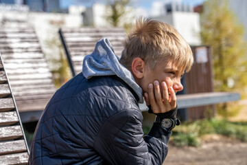 Teen boy sits on a weathered wooden bench, covering his face with his hands, projecting a sense of...