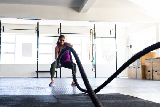Dedicated biracial young woman training with battle ropes in health club against window, copy space