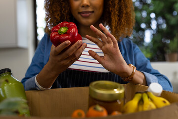 Happy biracial woman unpacking grocery shopping from box in kitchen holding red pepper