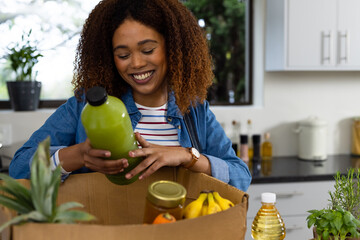 Happy biracial woman unpacking grocery shopping from recycled box in kitchen