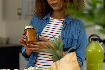 Midsection of biracial woman unpacking grocery shopping holding jar in kitchen holding bananas