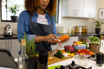 Focused biracial woman preparing vegetables in kitchen peeling carrot
