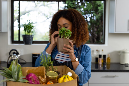 Happy biracial woman unpacking grocery shopping from box in kitchen and smelling herbs - Powered by Adobe