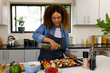 Happy biracial woman in apron preparing food in kitchen pouring oil on vegetables